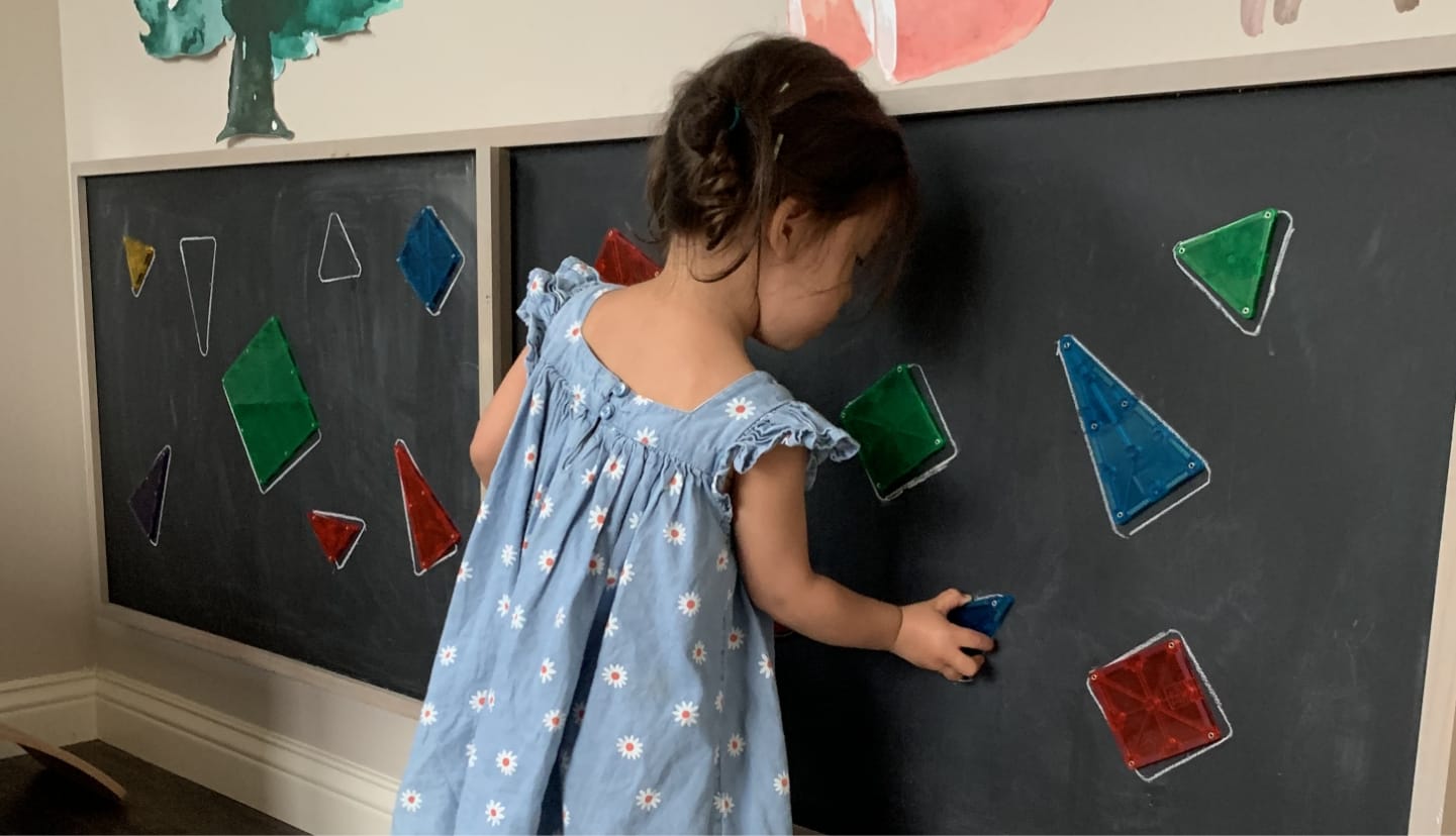 Young girl matching MAGNA-TILES® squares and triangles in hand-drawn shapes on a chalkboard
